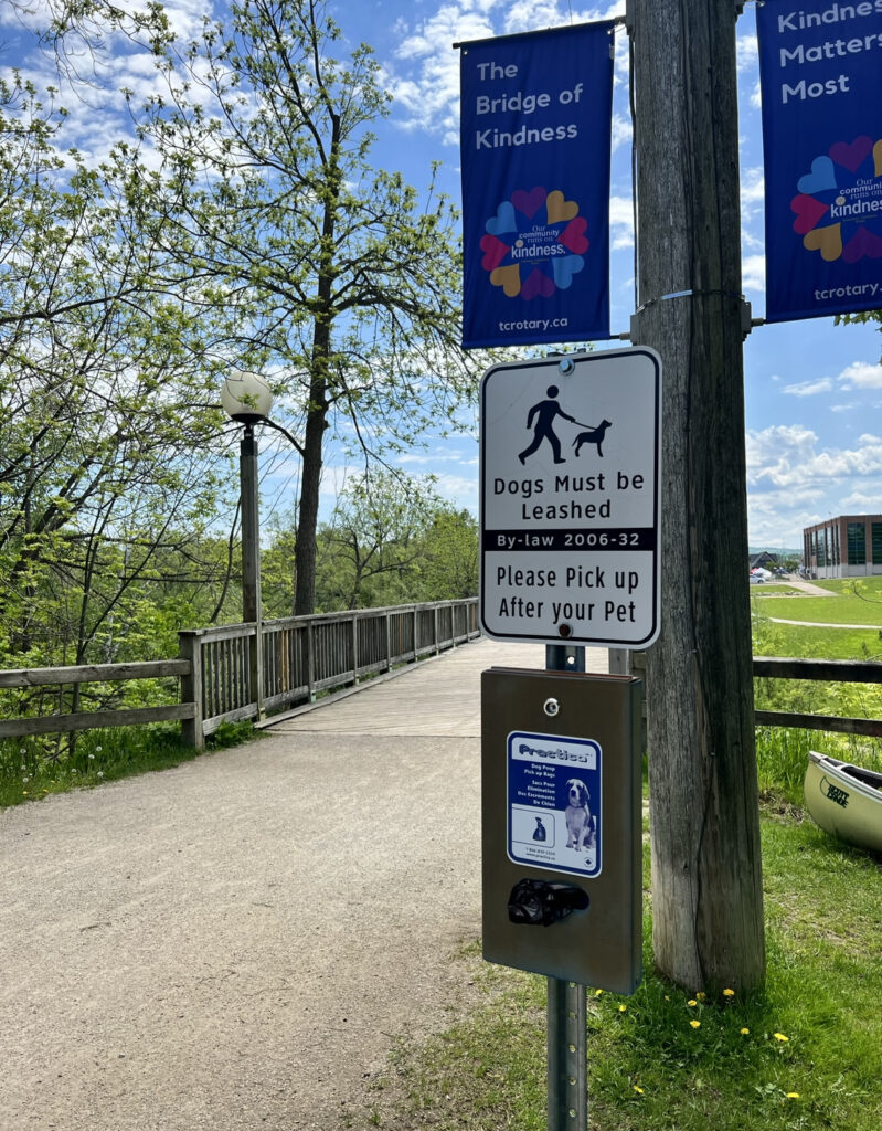 The Georgian Trail The Bridge of Kindness in Thornbury. Sign states Dogs must be leashed, please pick up after your pet (scoop 2 of course) and there are pet waste bags in the image near the bridge.